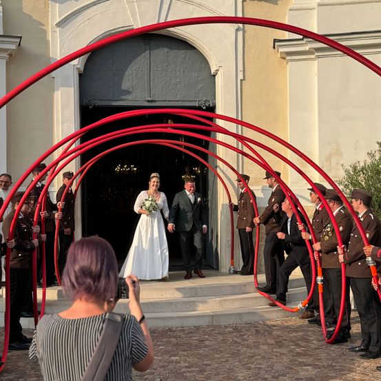 Ein frisch verheiratetes Paar geht durch einen roten Bogen, während Militärangehörige rote Röhren halten. Ein Fotograf hält den Moment fest.