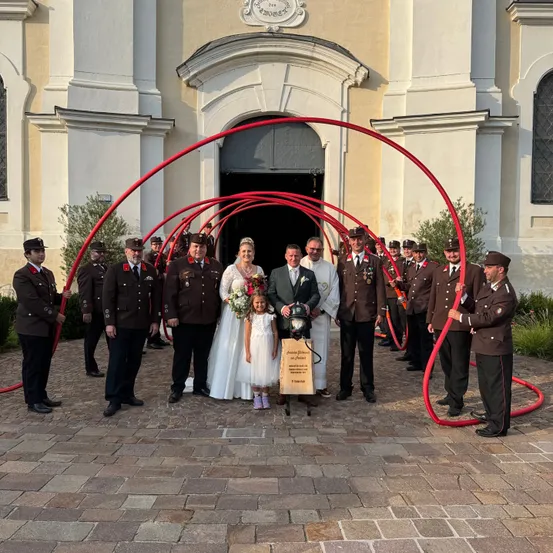 Eine Hochzeitsgesellschaft steht vor einer Kirche mit roten Bögen. Die Braut und der Bräutigam tragen Hochzeitskleidung. Hinter ihnen halten Soldaten in Uniform rote Objekte, möglicherweise Schläuche.