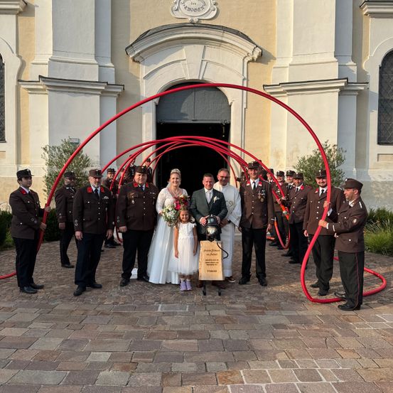 Eine Hochzeitsgesellschaft steht vor einer Kirche mit roten Bögen. Die Braut und der Bräutigam tragen Hochzeitskleidung. Hinter ihnen halten Soldaten in Uniform rote Objekte, möglicherweise Schläuche.