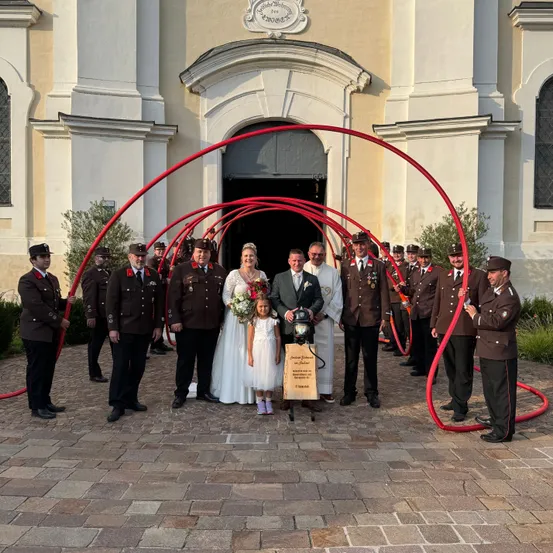Eine Hochzeitsgesellschaft steht vor einer Kirche mit Feuerwehrleuten. Das Brautpaar wird von Feuerwehrleuten umringt, die einen Feuerlöscher halten.