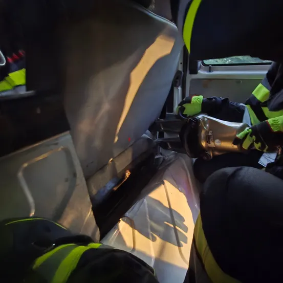Ein Feuerwehrmann arbeitet im Inneren eines Fahrzeugs und benutzt ein Werkzeug. Der Innenraum ist beleuchtet, mit einer Plastiktüte auf dem Boden. Eine weitere Person steht in der Nähe.