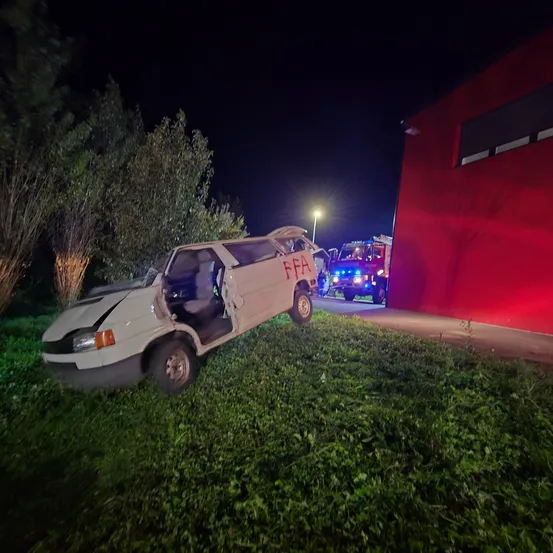 Ein weißer Van ist nachts gegen ein rotes Gebäude gekracht. Die Tür des Vans ist offen und die Front ist beschädigt. Ein Feuerwehrwagen ist in der Nähe geparkt, mit blinkenden Lichtern.