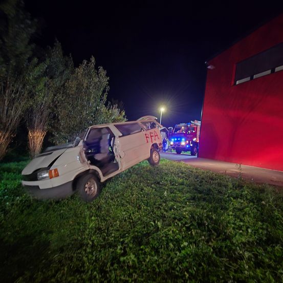 Ein weißer Van ist nachts gegen ein rotes Gebäude gekracht. Die Tür des Vans ist offen und die Front ist beschädigt. Ein Feuerwehrwagen ist in der Nähe geparkt, mit blinkenden Lichtern.