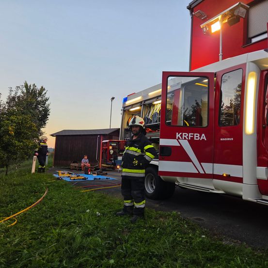 Ein Feuerwehrmann steht neben einem roten Feuerwehrauto mit offener Tür. Ein anderer Feuerwehrmann sitzt in der Nähe. Ein Gebäude mit roter Wand ist im Hintergrund.