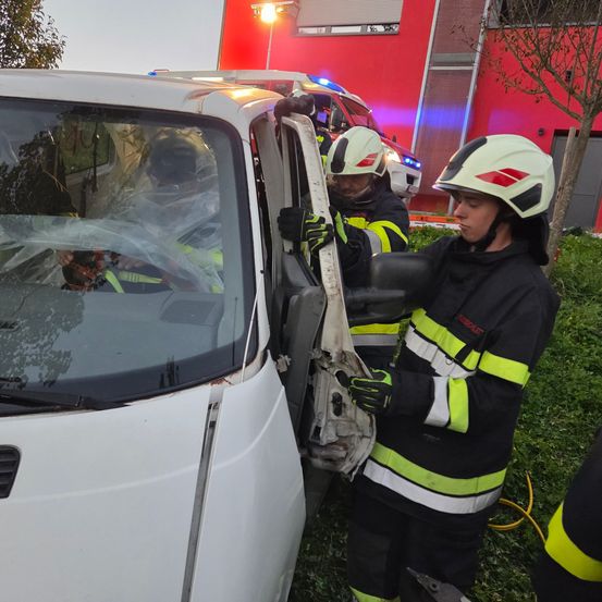 Zwei Feuerwehrleute öffnen die Tür eines weißen Lieferwagens. Die Windschutzscheibe des Wagens ist zerbrochen. Sie tragen Helme und Handschuhe. Ein Polizeiauto ist hinter ihnen geparkt.
