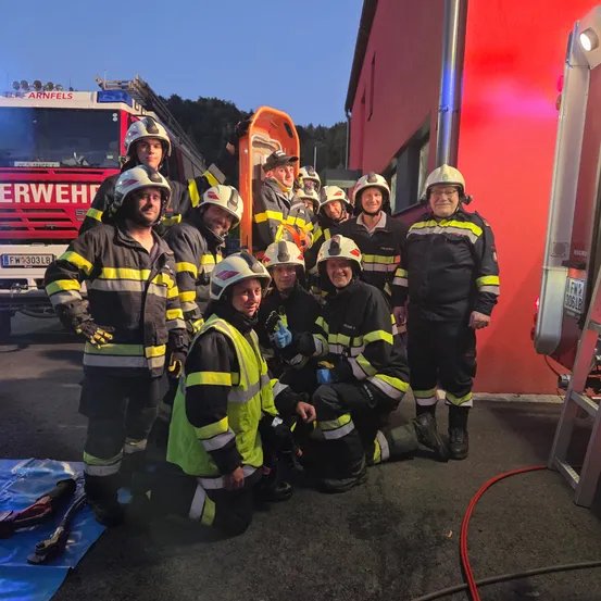 Eine Gruppe von Feuerwehrleuten posiert für ein Foto vor einer roten Feuerwache mit Ausrüstung im Hintergrund.