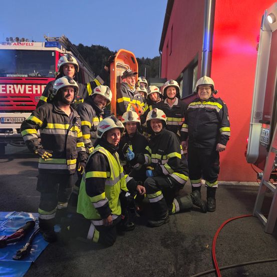 Eine Gruppe von Feuerwehrleuten posiert für ein Foto vor einer roten Feuerwache mit Ausrüstung im Hintergrund.
