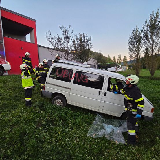 Feuerwehrleute helfen einem beschädigten weißen Van mit Graffiti in der Nähe eines roten Gebäudes auf einem Rasen mit Bäumen und einem geparkten weißen Auto.