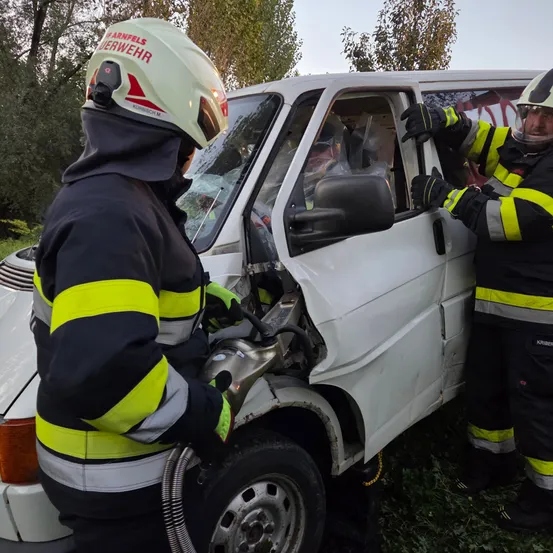 Zwei Feuerwehrleute in Schutzkleidung arbeiten daran, die Tür eines beschädigten weißen Lieferwagens zu öffnen. Der Schauplatz ist im Freien mit Bäumen im Hintergrund.