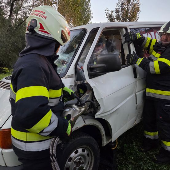 Zwei Feuerwehrleute in Schutzkleidung arbeiten daran, die Tür eines beschädigten weißen Lieferwagens zu öffnen. Der Schauplatz ist im Freien mit Bäumen im Hintergrund.