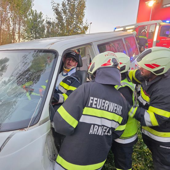 Drei Feuerwehrleute helfen einem Mann in einem Van. Er hat eine Halskrause und scheint verletzt zu sein. Sie befinden sich in einem Grasbereich mit Bäumen und einem Feuerwehrwagen im Hintergrund.