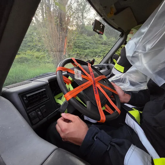 Eine Person in einer Sicherheitsweste sitzt in einem Fahrzeug und hält das Lenkrad fest, das mit orangefarbenen Bändern umwickelt ist. Der Armaturenbrett ist sichtbar, und der Innenraum des Fahrzeugs zeigt einen Rückspiegel und eine durchsichtige Plastikabdeckung über dem vorderen Fenster.