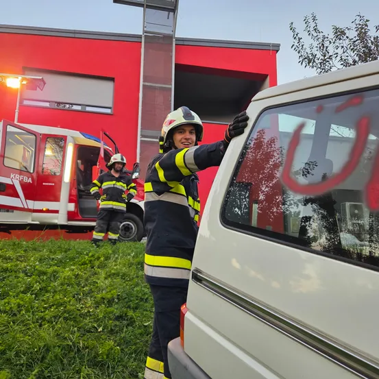 Ein Feuerwehrmann mit Helm steht neben einem Van mit Graffiti vor einem roten Gebäude. Ein weiterer Feuerwehrmann steht neben einem Feuerwehrwagen mit offener Tür.