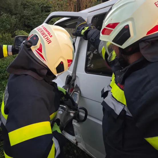 Zwei Feuerwehrleute in Schutzkleidung arbeiten an einem Fahrzeug. Einer benutzt ein Werkzeug, um die Tür zu öffnen, während der andere zusieht. Das Fahrzeug ist weiß mit roten und gelben Markierungen.