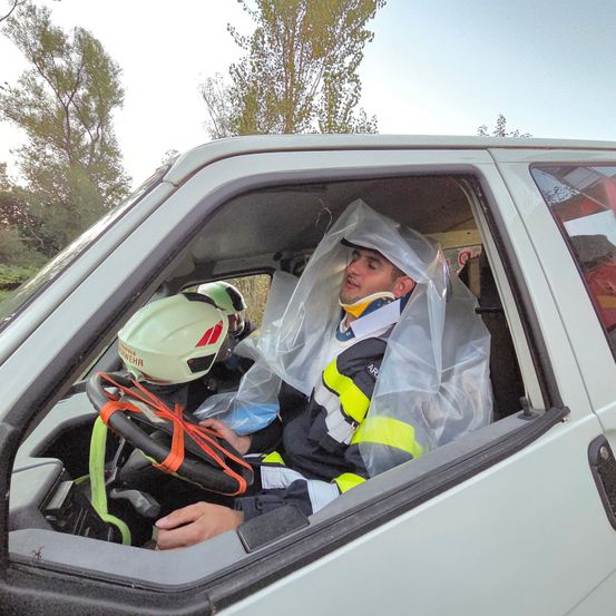 Ein Mann mit einem Nackenschutz sitzt auf dem Fahrersitz eines weißen Fahrzeugs, im Hintergrund befinden sich zwei Personen mit Helmen.