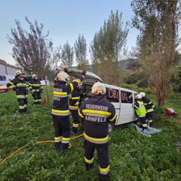 Eine Gruppe Feuerwehrleute in Schutzkleidung arbeitet daran, jemanden aus einem weißen Van zu retten, der auf seiner Seite in einem Grasfeld umgekippt ist. Bäume und ein Gebäude sind im Hintergrund zu sehen.