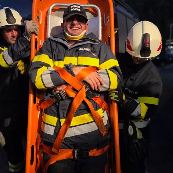Ein Feuerwehrmann mit Halskralle wird von zwei weiteren Personen unterstützt, die Helme und Handschuhe tragen. Sie befinden sich auf einer orangefarbenen Trage, wahrscheinlich in der Nähe eines Feuerwehrwagens.