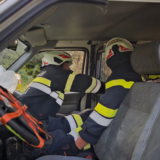 Zwei Feuerwehrleute in voller Schutzausrüstung sitzen auf dem Fahrer- und Beifahrersitz eines Fahrzeugs. Sie tragen beide Helme mit dem Wort Feuerwehr.
