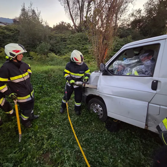 Drei Feuerwehrleute sind auf einem Grasfeld mit einem weißen Lieferwagen. Einer benutzt ein Elektrowerkzeug an der Seite des Wagens. Der Fahrer ist drinnen, und ein anderer Feuerwehrmann ist in der Nähe.