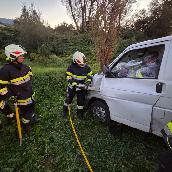 Drei Feuerwehrleute sind auf einem Grasfeld mit einem weißen Lieferwagen. Einer benutzt ein Elektrowerkzeug an der Seite des Wagens. Der Fahrer ist drinnen, und ein anderer Feuerwehrmann ist in der Nähe.