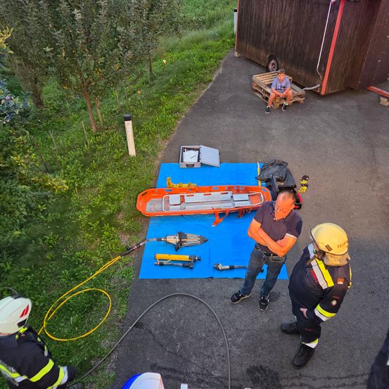 Einige Feuerwehrleute sind um eine orangefarbene Trage auf einem blauen Teppich versammelt. Ein Feuerwehrmann beobachtet, während die anderen beiden in ein Gespräch vertieft sind. In der Nähe sitzt ein Junge auf einer Holzpalette, und im Hintergrund befindet sich ein kleines Häuschen.