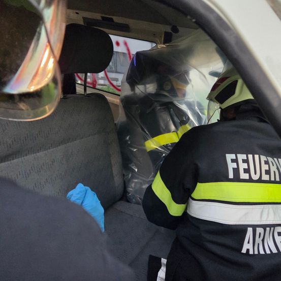 Zwei Feuerwehrleute in Schutzkleidung sind in einem Fahrzeug, einer trägt einen blauen Handschuh. Das Fahrzeug hat graue Sitze und eine transparente Plastikbarriere.