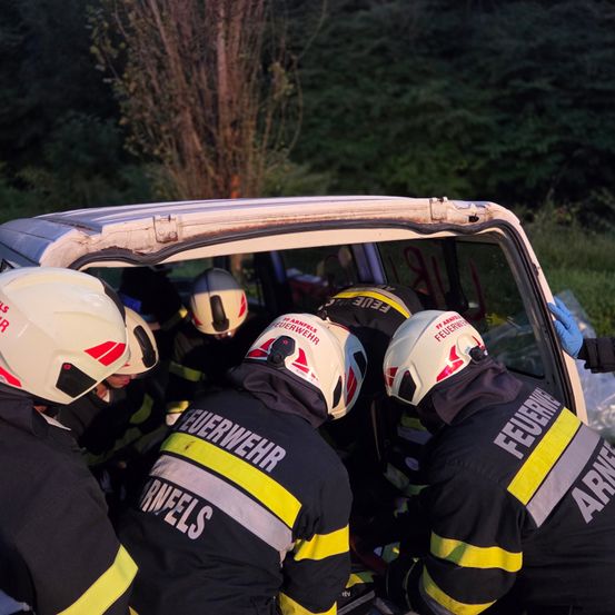 Feuerwehrleute in Helmen kümmern sich um ein Unfallfahrzeug. Das Auto weist sichtbare Schäden auf.