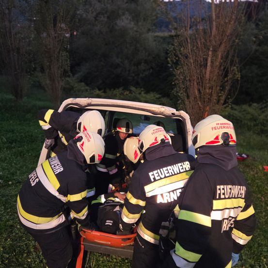 Eine Gruppe von Feuerwehrleuten in Uniform kümmert sich um eine verletzte Person auf einer Trage in einem Grasbereich.