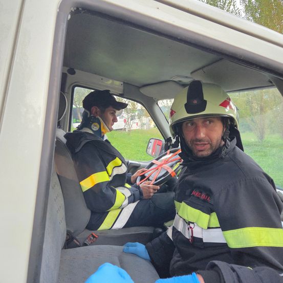 Zwei Feuerwehrleute sitzen in einem Fahrzeug. Einer trägt eine Halskrause und der andere trägt einen Helm und Handschuhe.
