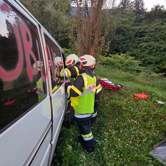 Feuerwehrleute in Schutzausrüstung stehen neben einem Fahrzeug mit Graffiti an der Seite, in der Nähe eines Baumes, mit Notfallgerät auf dem Boden.