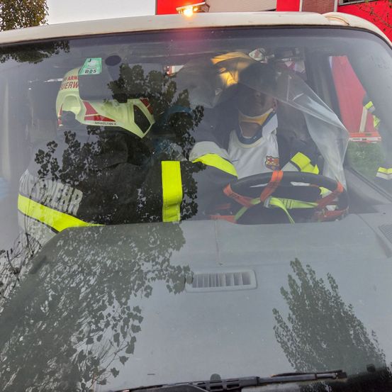 Eine Person mit Halskralle sitzt in einem Auto mit Feuerwehrleuten. Die Windschutzscheibe spiegelt Bäume und ein Gebäude wider.