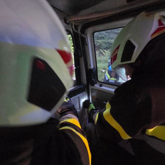 Feuerwehrleute in einem Fahrzeug mit Helmen, die aus dem Fenster schauen auf eine Grasfläche und Bäume. Eine Person in einer gelben Weste ist draußen sichtbar.