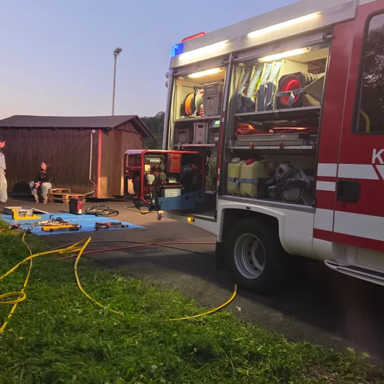Ein Feuerwehrfahrzeug mit offenen Türen steht auf einem Parkplatz. Im Inneren des Fahrzeugs ist Ausrüstung sichtbar. Zwei Personen sitzen in der Nähe auf einem Palette.