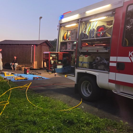 Ein Feuerwehrfahrzeug mit offenen Türen steht auf einem Parkplatz. Im Inneren des Fahrzeugs ist Ausrüstung sichtbar. Zwei Personen sitzen in der Nähe auf einem Palette.
