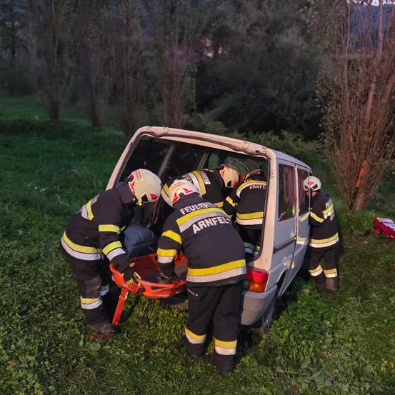 Eine Gruppe Feuerwehrleute steht neben einem beschädigten Van und bereitet sich darauf vor, eine Person mit einer Trage herauszuholen. Sie tragen vollständige Schutzausrüstung.