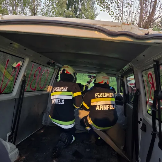 Zwei Feuerwehrleute sitzen in einem Van, einer kniet und der andere sitzt, beide tragen Uniformen und Helme. Der Van hat Graffiti auf den Fenstern.