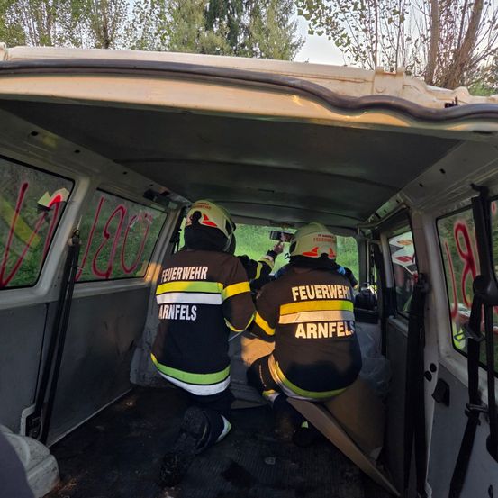 Zwei Feuerwehrleute sitzen in einem Van, einer kniet und der andere sitzt, beide tragen Uniformen und Helme. Der Van hat Graffiti auf den Fenstern.