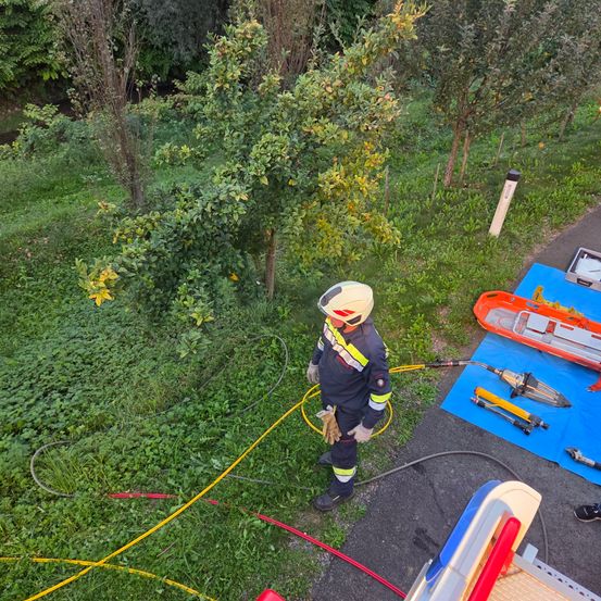 Ein Feuerwehrmann steht auf Gras mit Ausrüstung um ihn herum. Werkzeuge und eine Trage liegen auf einem blauen Tarp. Bäume und Pflanzen umgeben das Gebiet.