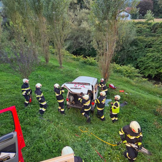 Eine Gruppe von Feuerwehrleuten ist um einen weißen Van mit beschädigter Front und offener Hecktür versammelt, der sich auf einem Grasfeld befindet. Sie tragen alle Helme und Uniformen, einer hält eine gelbe Seile.
