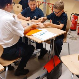 Zwei junge Jungen in Uniformen sitzen an einem Schreibtisch mit einer erwachsenen Frau, die ihre Hände hält, während sie etwas bearbeiten. Ein Feuerlöscher und andere Ausrüstung sind im Hintergrund.