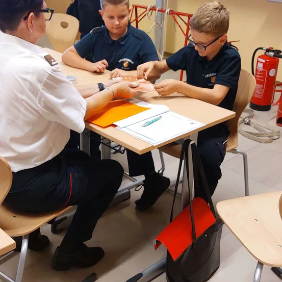 Zwei Jungen und eine Frau in Uniform sitzen an einem Tisch, wahrscheinlich in einem Klassenzimmer. Die Jungen tragen Uniformen, einer hält ein Stück Papier, während der andere schreibt. Ein Feuerlöscher ist im Hintergrund zu sehen.