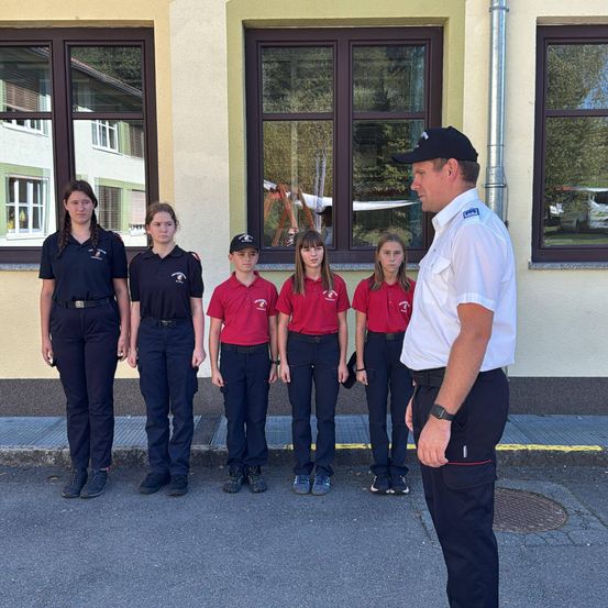 Eine Gruppe junger Mädchen und Jungen in roten Hemden steht in einer Reihe mit einem Mann in weißer Uniform vor einem Gebäude.