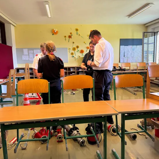 Ein Klassenzimmer mit verstreuten Tischen und Stühlen, einem Mann und zwei Frauen in Uniformen sowie Feuerlöschern und Werkzeugen auf dem Boden.