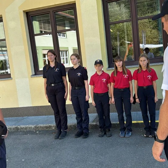 Fünf Mädchen und ein Junge in roten Uniformen stehen in einer Reihe vor einem Gebäude mit Fenstern. Sie gehören wahrscheinlich zu einer Jugendgruppe.