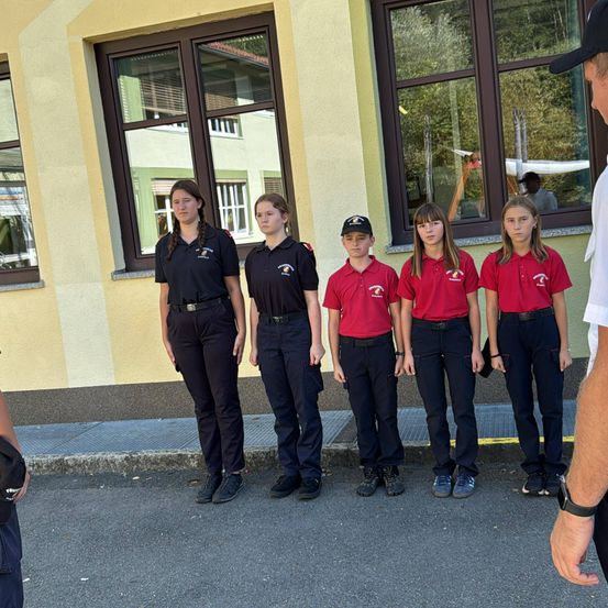 Fünf Mädchen und ein Junge in roten Uniformen stehen in einer Reihe vor einem Gebäude mit Fenstern. Sie gehören wahrscheinlich zu einer Jugendgruppe.