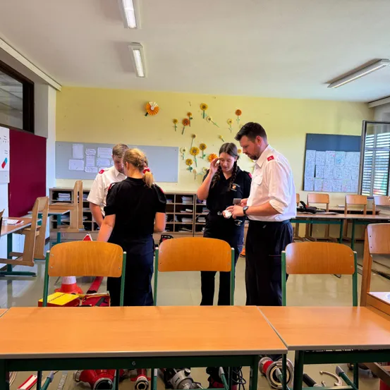 Eine Gruppe von Menschen in einem Klassenzimmer, drei von ihnen stehend und einer telefoniert. Zwei Tische stehen im Vordergrund und verschiedene Geräte sind auf dem Boden abgestellt.