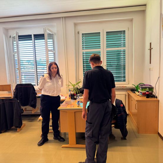 Eine Frau und ein Mann stehen in einem Büro und sehen sich gegenüber. Sie trägt eine Brille und eine Uniform, während er ein schwarzes Shirt trägt. Ein Schreibtisch und Pflanzen befinden sich zwischen ihnen.