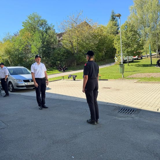 Drei uniformierte Personen stehen auf einem Parkplatz. Einer schaut in die Kamera, die anderen beiden schauen einander an. Ein Auto ist in der Nähe geparkt. Bäume und ein Grasbereich sind im Hintergrund.