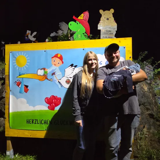 Ein Mann und eine Frau stehen neben einem bunten Schild mit Cartoon-Figuren, halten ein Baby und lächeln. Das Schild sagt 'Herzlichen Gluckwunsch' mit einem Storch und anderen Illustrationen.