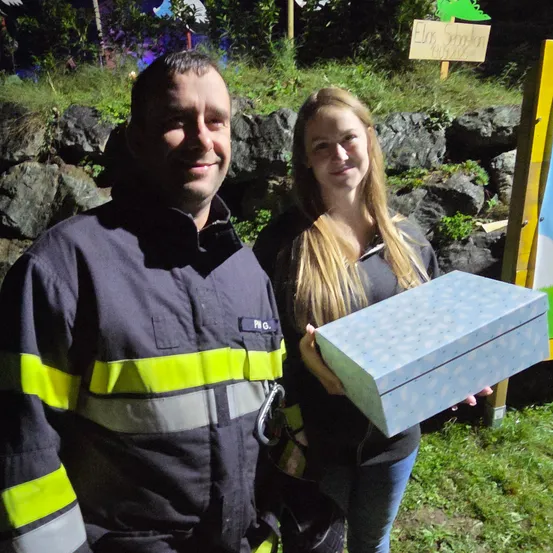 Ein Feuerwehrmann und eine Frau stehen lächelnd vor einer Steinmauer. Die Frau hält eine blaue Schuhschachtel. Ein Schild ist an der Wand befestigt.